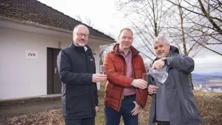 Drei Männer in Jacken stehen lächelnd vor einem Haus. Der Mann auf der rechten Seite gießt Wasser in ein Glas, das der Mann in der Mitte hält. Der Mann auf der linken Seite hält ein Glas.