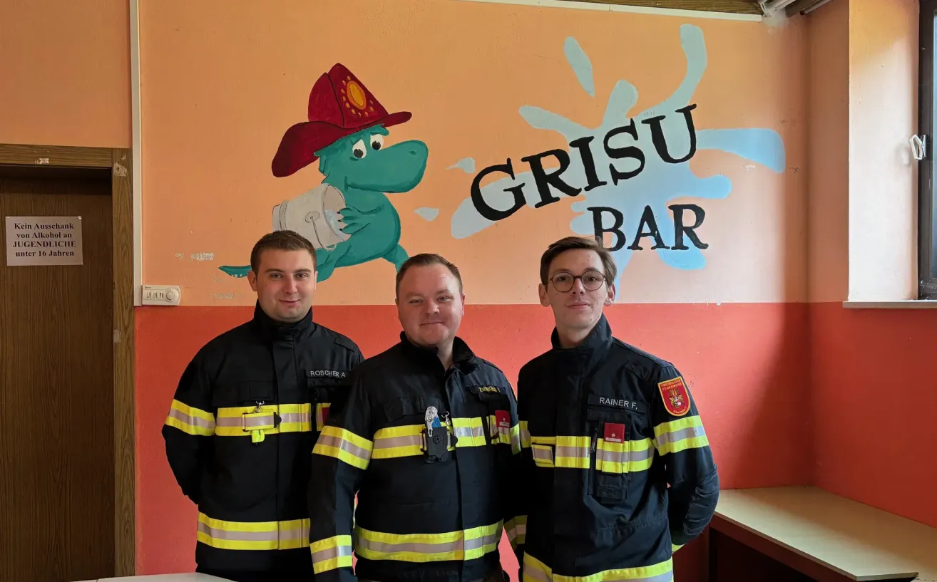 Drei Feuerwehrleute stehen vor einer Wand mit dem Wort Grisu Bar und einem Dinosaurier. Einer hat einen roten Aufnäher am Arm.
