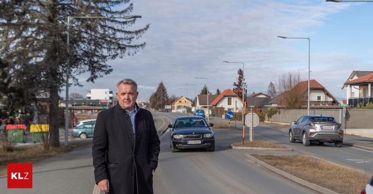 Ein Mann steht am Straßenrand neben einem geparkten Auto, mit Häusern und Bäumen im Hintergrund.