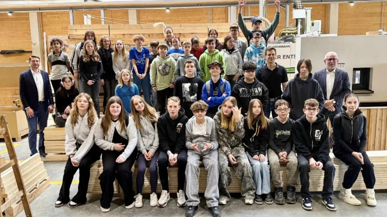 Eine Gruppe von Schülern posiert für ein Foto in einer Werkstatt. Sie sitzen und stehen in einer Reihe auf einer Holzbank. Die Schüler sind lässig gekleidet. Der Hintergrund zeigt Holzkonstruktionen und Ausrüstung.
