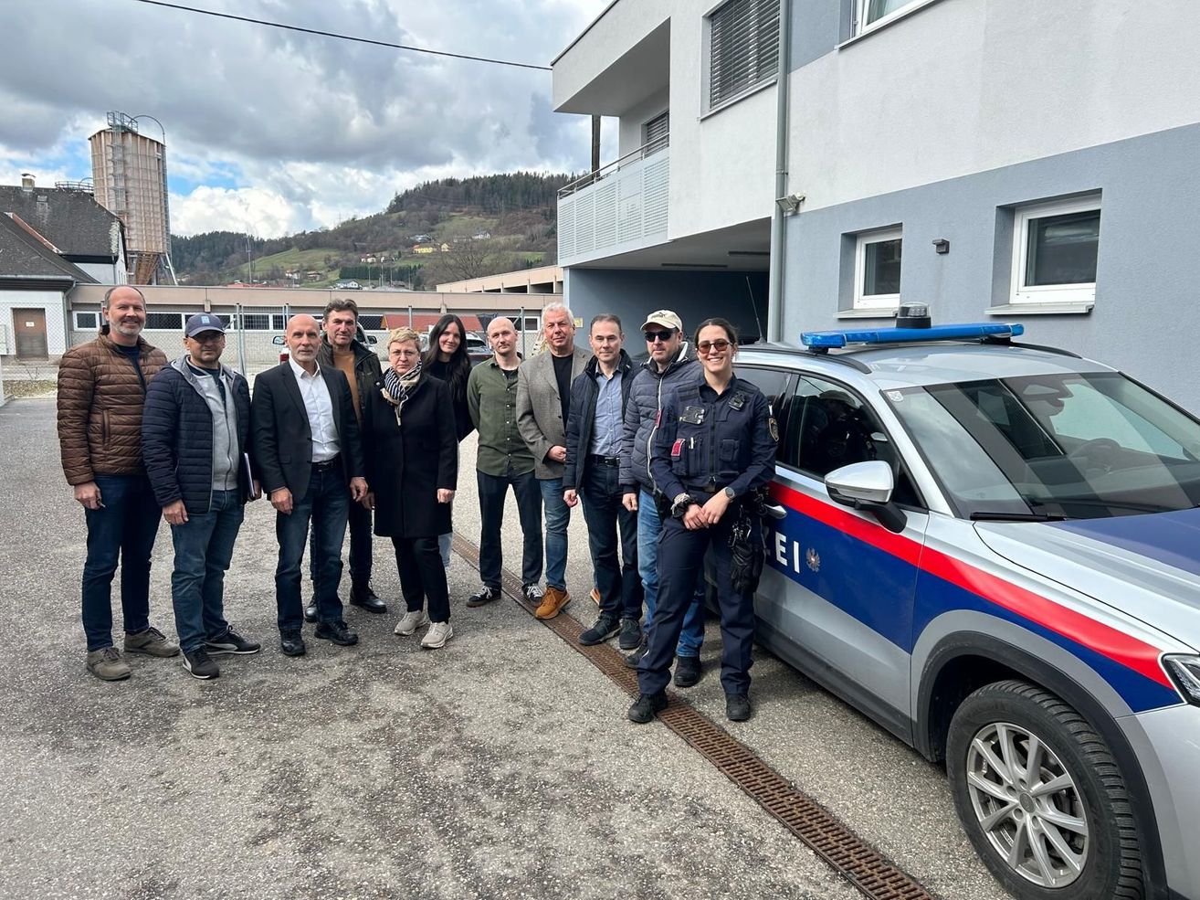 Eine Gruppe von Menschen posiert für ein Foto vor einem Polizeiauto. Sie stehen auf einer gepflasterten Fläche vor einem Gebäude.