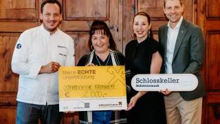 Vier Personen posieren für ein Foto, wobei eine einen Scheck über 2.000 Euro hält. Die Frau hält ein Schild für die Österreichische Krebshilfe. Der Mann trägt eine Kochuniform. Der Mann rechts hält ein Schild für Schlosskeller Südsteiermark.