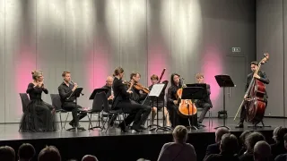 Ein Konzert auf einer Bühne mit Musikern, die verschiedene Instrumente spielen. Das Publikum sitzt vor den Darstellern. Die Bühne hat einen einfachen Hintergrund.