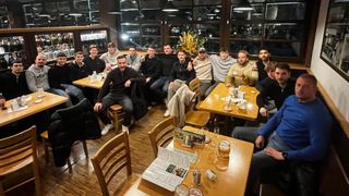 Eine Gruppe von Männern sitzt in einem Restaurant um einen Tisch herum, einige halten Getränke. Sie scheinen Freunde zu sein, wobei mehrere ähnliche Kleidung tragen. Der Tisch hat Biergläser und eine Speisekarte.