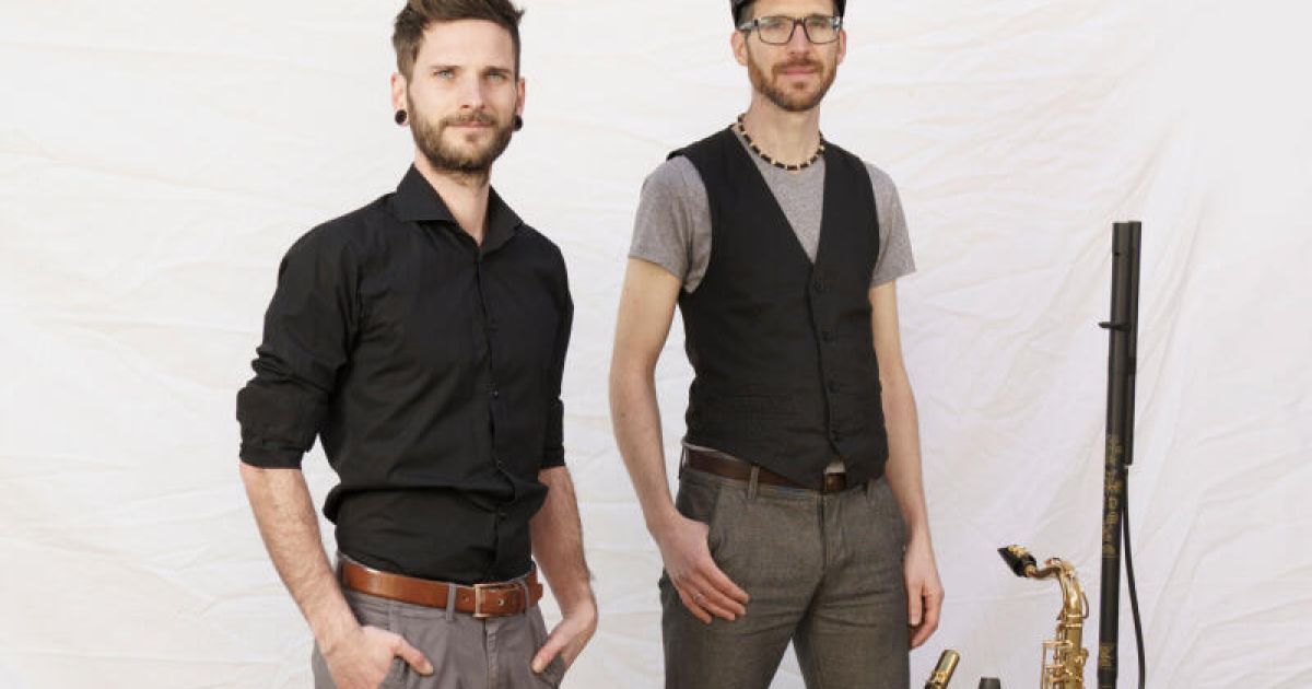 Zwei Männer stehen nebeneinander vor einem weißen Hintergrund. Einer trägt ein schwarzes Hemd, der andere ein graues T-Shirt und eine schwarze Weste. Beide haben Bärte.