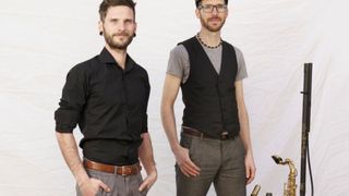 Zwei Männer stehen nebeneinander vor einem weißen Hintergrund. Einer trägt ein schwarzes Hemd, der andere ein graues T-Shirt und eine schwarze Weste. Beide haben Bärte.