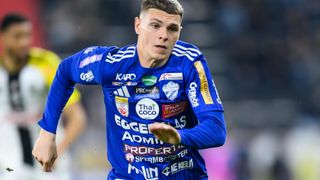 Ein Fußballspieler läuft auf dem Feld in einem blauen Trikot mit mehreren Werbungen. Sein Gesichtsausdruck ist konzentriert.