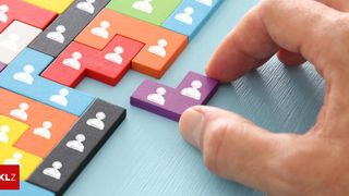Eine Hand platziert ein lila Puzzleteil mit weißen Symbolen in ein buntes Puzzlebrett.