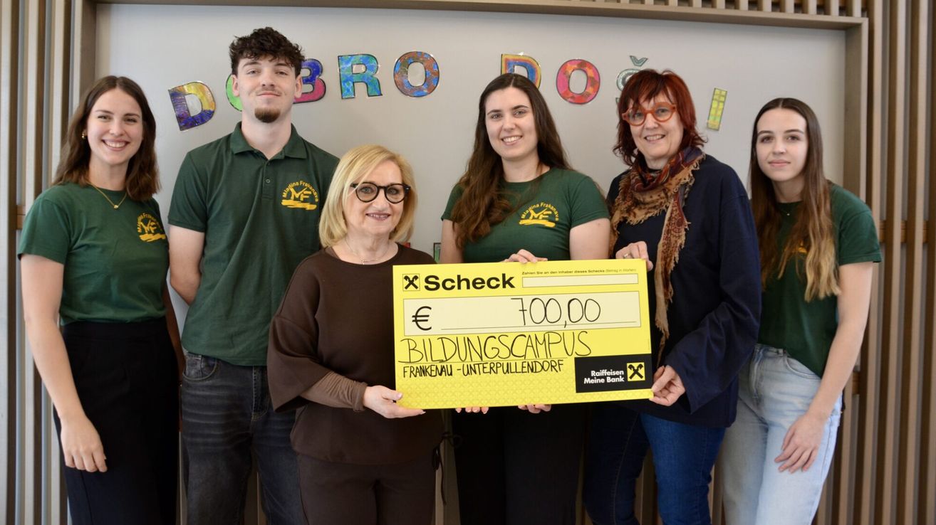 Fünf Personen stehen vor einem farbenfrohen Hintergrund. Die Frau auf der linken Seite hält einen gelben Scheck über 700 Euro. Hinter ihr hält eine weitere Frau einen Scheck. Die anderen tragen grüne Shirts mit einem Logo.