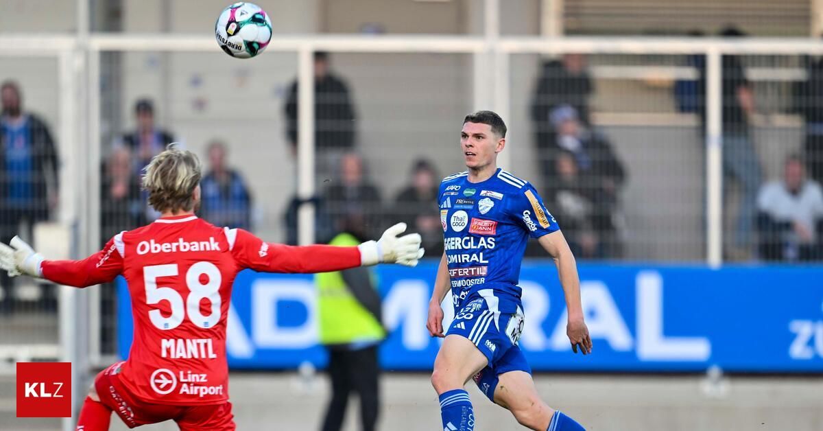 Ein Fußballspieler in Blau kopfballt den Ball, während ein anderer Spieler in Rot zusieht. Im Hintergrund ist eine verschwommene Menge von Zuschauern zu sehen, die das Spiel beobachten.