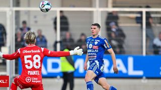 Ein Fußballspieler in Blau kopfballt den Ball, während ein anderer Spieler in Rot zusieht. Im Hintergrund ist eine verschwommene Menge von Zuschauern zu sehen, die das Spiel beobachten.