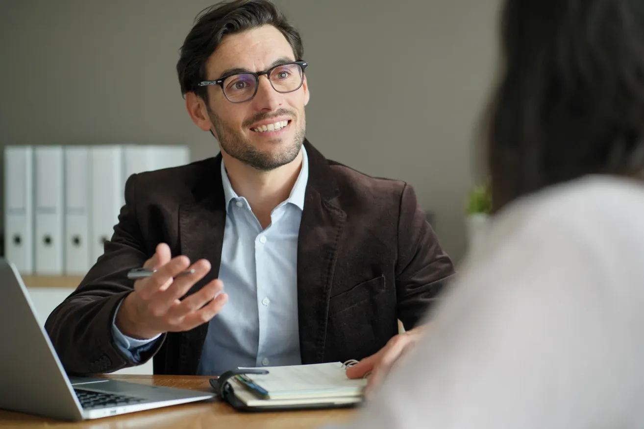 Ein Mann mit Brille im Anzug sitzt an einem Schreibtisch, hält einen Stift in der Hand und lächelt, während er mit einer anderen Person spricht.