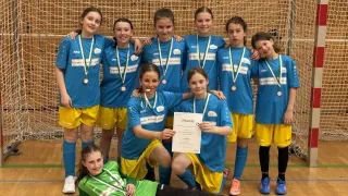 Eine Gruppe junger Mädchen in blauen und gelben Trikots steht vor einem Netz. Ein Mädchen hält ein Zertifikat. Sie tragen alle Medaillen.