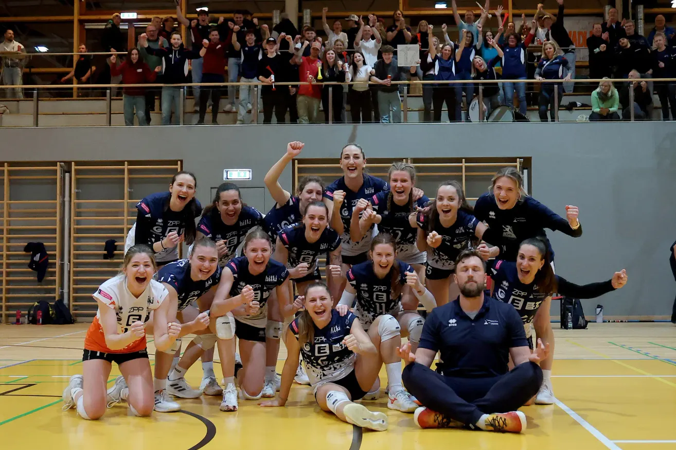 Ein Volleyballteam feiert einen Sieg auf einem Spielfeld mit einem Trainer vorne. Fans jubeln von den Tribünen.
