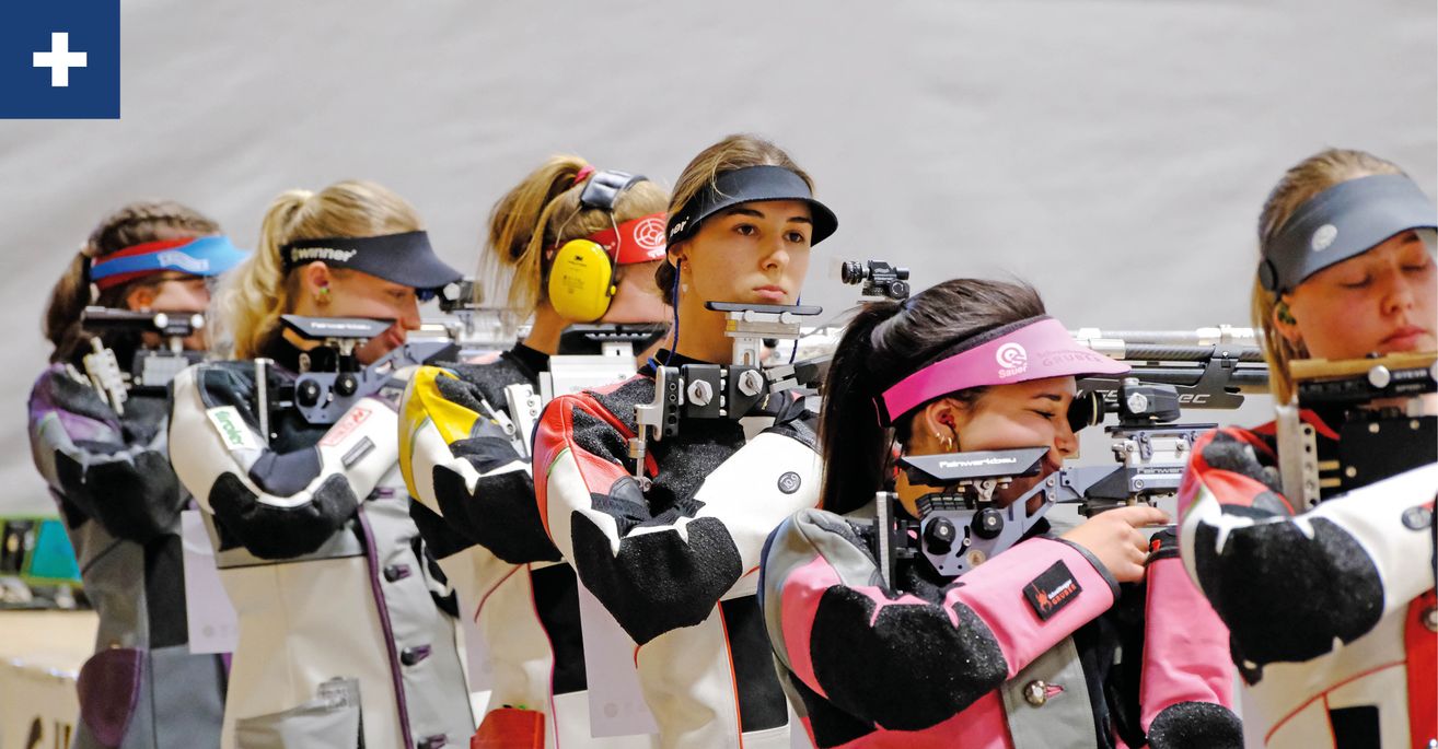 Fünf Frauen, alle in Schießjacken und Stirnbändern gekleidet, zielen mit ihren Gewehren auf ein Ziel. Sie stehen in einer Reihe und tragen alle Gehörschutz.