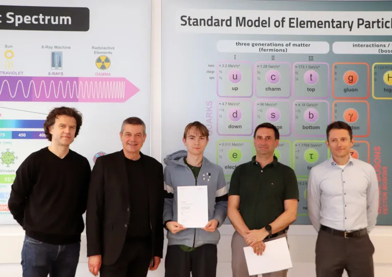 Fünf Männer stehen vor einem Schild mit der Aufschrift 'Standardmodell der Elemente.' Sie halten Papiere in der Hand. Der mittlere Mann hält ein weißes Papier mit gedrucktem Text.