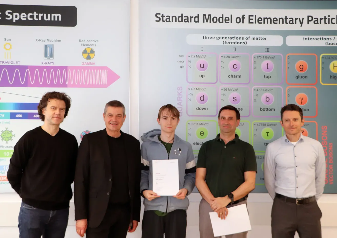 Fünf Männer stehen vor einem Schild mit der Aufschrift 'Standardmodell der Elemente.' Sie halten Papiere in der Hand. Der mittlere Mann hält ein weißes Papier mit gedrucktem Text.