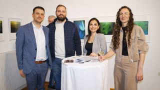 Vier lächelnde Personen stehen in der Nähe eines Tisches mit Papieren und einem Weinglas. Dahinter befinden sich Kunstwerke an der Wand und ein weiterer Mann.