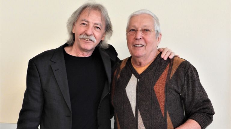 Zwei ältere Männer stehen nebeneinander. Ein Mann hat einen Schnurrbart, der andere trägt eine Brille. Beide lächeln.