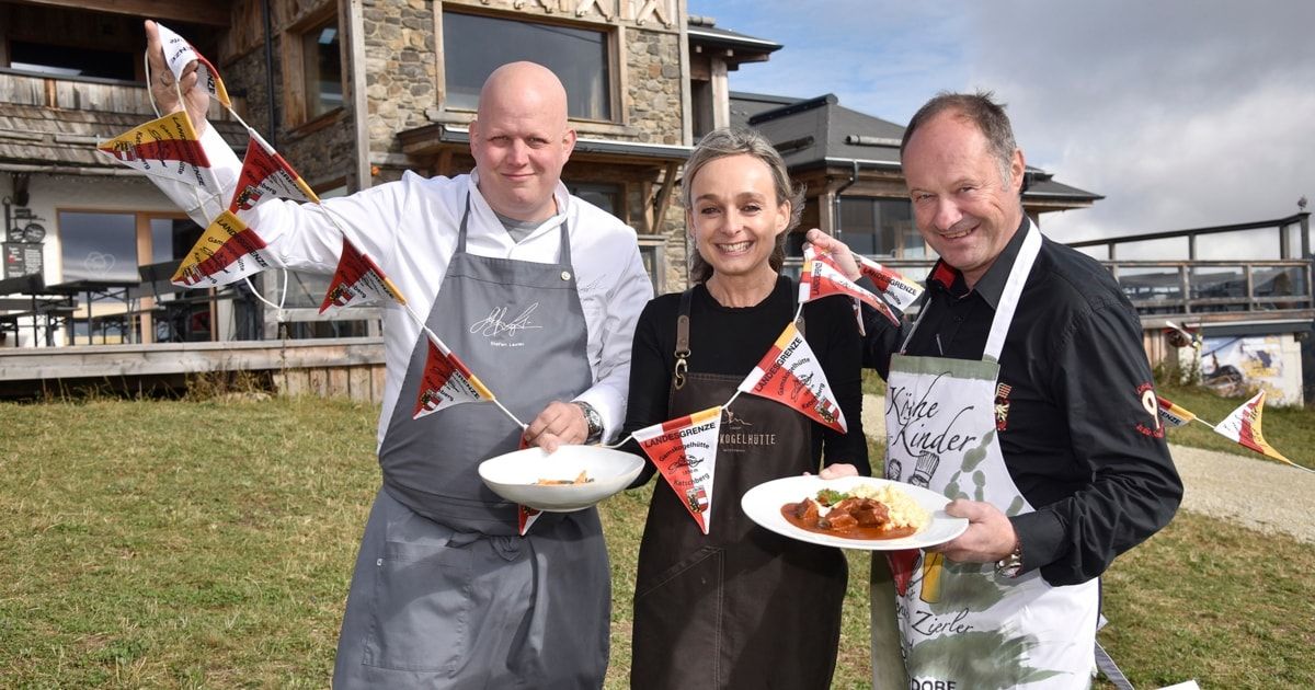 Drei Köche stehen mit Tellern voller Essen vor einem Steingebäude und lächeln in ihren Schürzen.