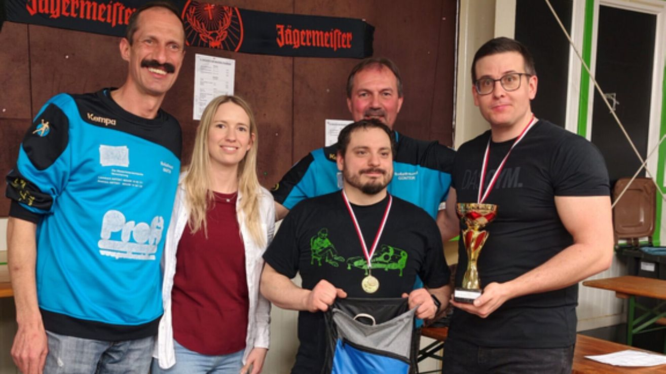 Eine Gruppe von Menschen lächelt und posiert für ein Foto mit Medaillen. Ein Mann hält eine Trophäe. Die Wand hinter ihnen hat ein Banner mit 'Jagermeister' darauf.