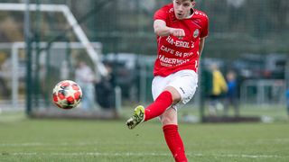 Ein Fußballspieler in einem roten Trikot tritt den Ball während eines Spiels. Der Ball ist in der Luft. Dahinter sind Zuschauer verschwommen.