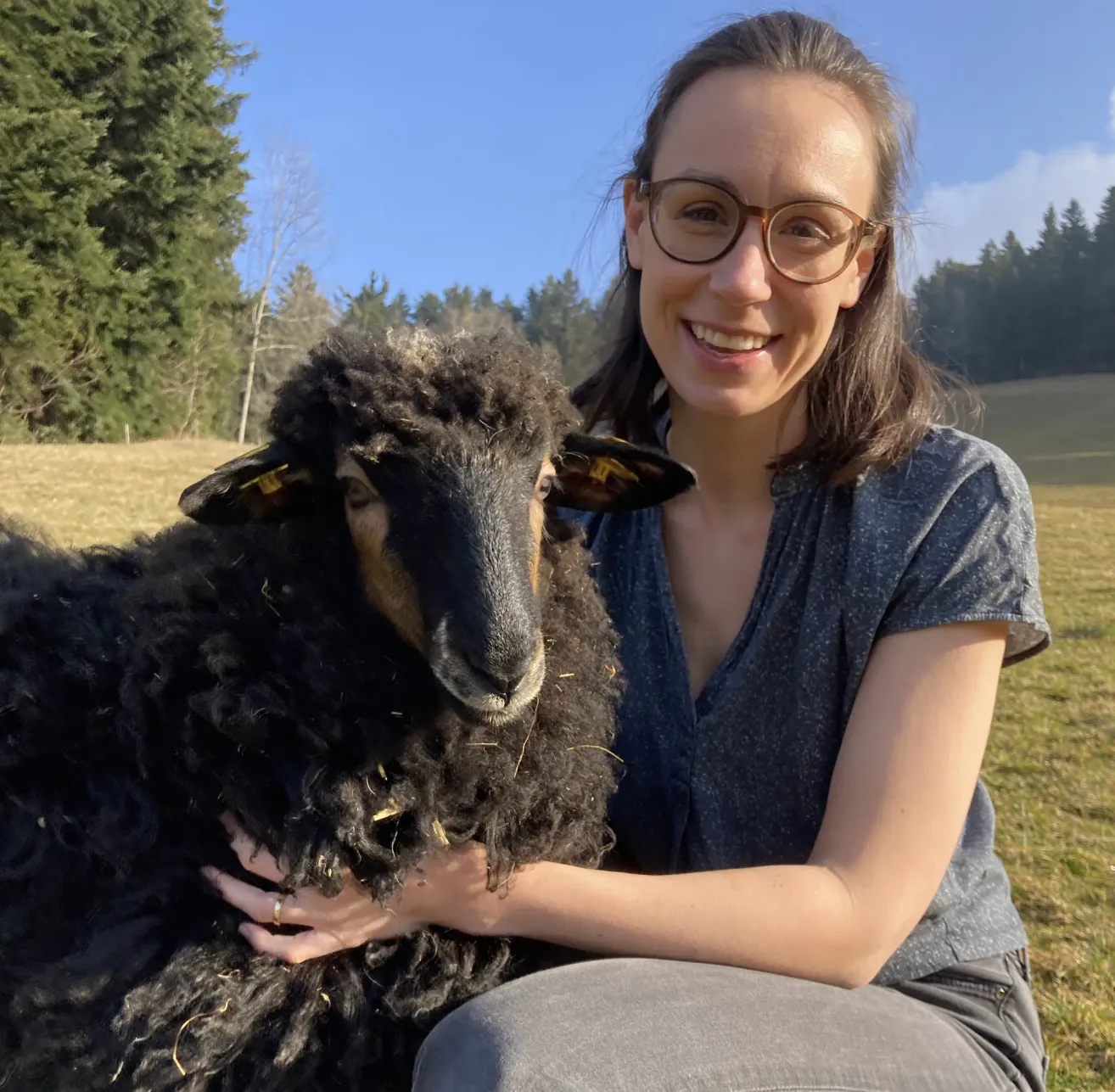 Eine Frau mit Brille hält ein schwarzes Schaf in einem Feld mit grünen Bäumen im Hintergrund.