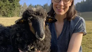 Eine Frau mit Brille hält ein schwarzes Schaf in einem Feld mit grünen Bäumen im Hintergrund.