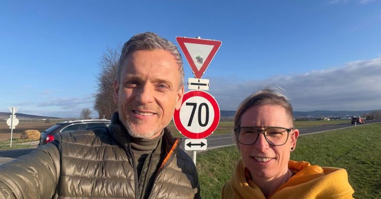 Zwei Personen stehen neben einer Straße mit einem Geschwindigkeitsbegrenzungsschild, das 70 km/h anzeigt. Der Mann trägt eine grüne Jacke, und die Frau trägt eine Brille und einen gelben Schal. Hinter ihnen befindet sich eine Rasenfläche und eine Straße mit einem Auto. Der Himmel ist blau mit einigen Wolken.