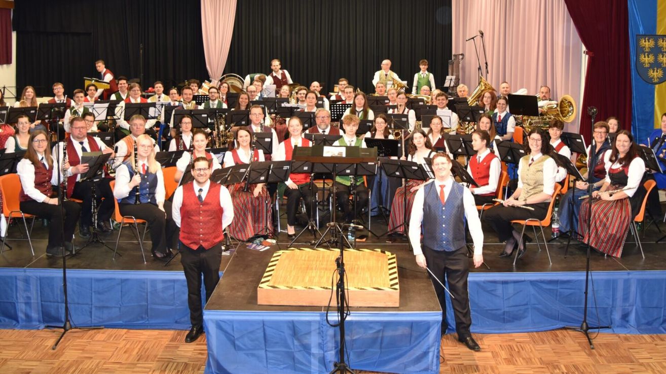 Ein großes Orchester posiert auf der Bühne mit einem Dirigenten vor einem Notenständer. Dahinter sitzen und stehen Musiker und spielen verschiedene Instrumente. Das Publikum sitzt im Hintergrund.