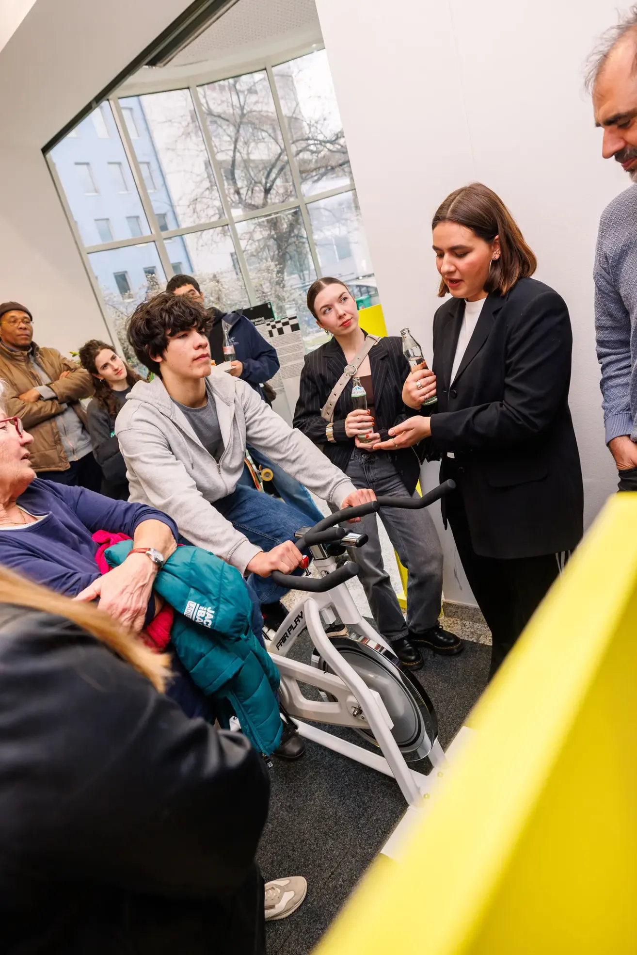 Eine Gruppe von Menschen versammelt sich um einen Mann auf einem stationären Fahrrad, wobei eine Frau eine Flasche vor ihm hält. Eine andere Frau hält eine Flasche und sie schauen alle auf ihn.