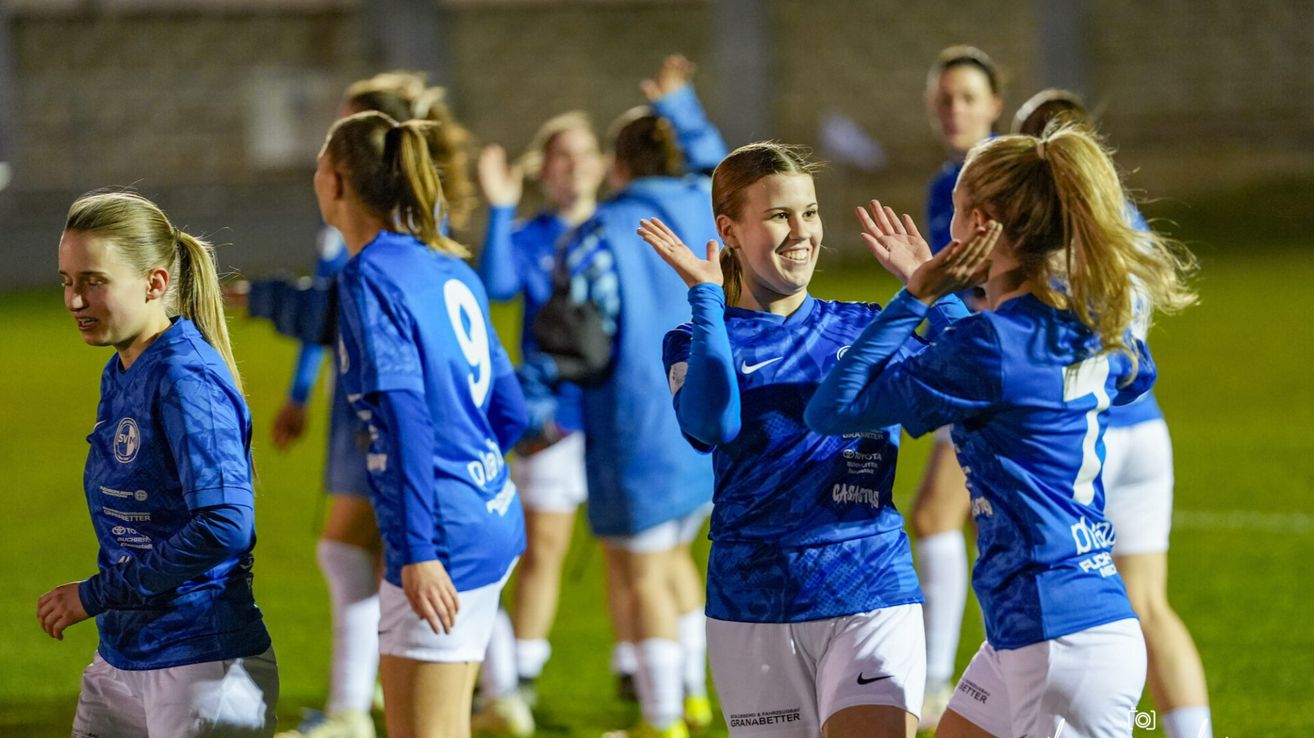 Eine Gruppe von Fußballerinnen in blauen Trikots feiert auf dem Spielfeld. Die Spielerin in der Mitte lächelt und andere klatschen mit den Händen.