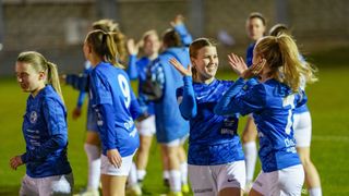 Eine Gruppe von Fußballerinnen in blauen Trikots feiert auf dem Spielfeld. Die Spielerin in der Mitte lächelt und andere klatschen mit den Händen.