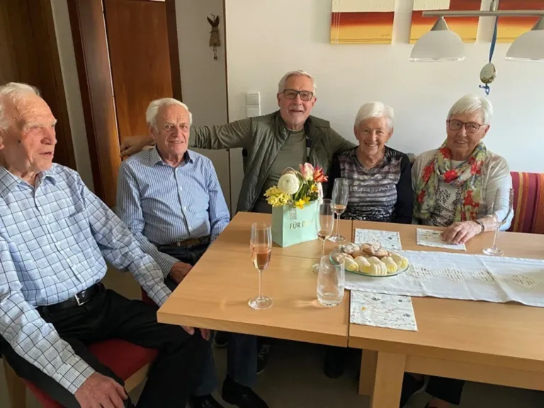 Five elderly individuals gather around a table, smiling, with a floral arrangement and wine glasses. A white tablecloth and napkins are on the table.