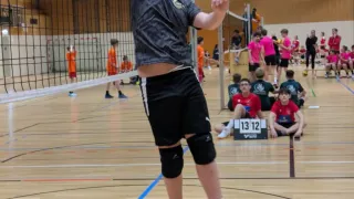 Ein Junge springt, um einen Volleyball in einer Halle zu schlagen. Zuschauer in pinken Trikots schauen zu, einige sitzen auf dem Boden. Der Platz hat farbige Markierungen.