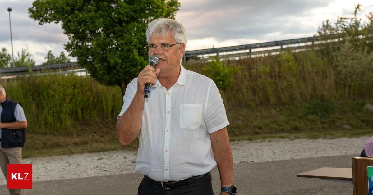 Ein älterer Mann mit weißen Haaren, der eine Brille, ein weißes Hemd und schwarze Hose trägt, steht im Freien und spricht in ein Mikrofon. Er befindet sich in einem Grasbereich mit einem Metallzaun und Bäumen im Hintergrund.