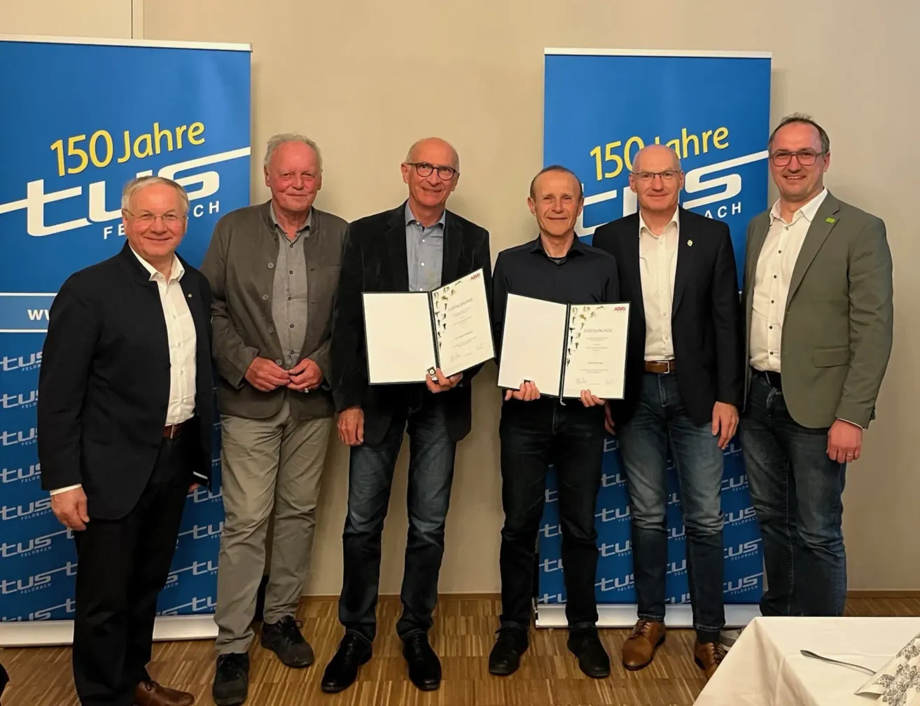 Fünf Männer stehen zusammen und halten Urkunden in den Händen. Sie sind in formeller Kleidung gekleidet. Einer der Männer trägt eine Brille und einen Gürtel. Hinter ihnen befinden sich zwei Banner mit Logos.