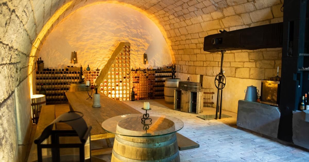 Ein Keller aus Stein mit einem Holztisch und Fässern mit Wein. Der Keller ist mit warmen Lichtern beleuchtet und verfügt über einen Projektor.