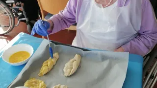 Eine ältere Frau im Rollstuhl malt mit einem Pinsel Teig in einer Küche. Es befinden sich vier Teigstücke auf dem Tablett.