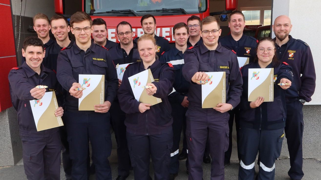 Eine Gruppe von Feuerwehrleuten posiert für ein Foto, alle in Uniform mit Namensschildern. Sie halten Urkunden und lächeln, stehen vor einem roten Feuerwehrauto.