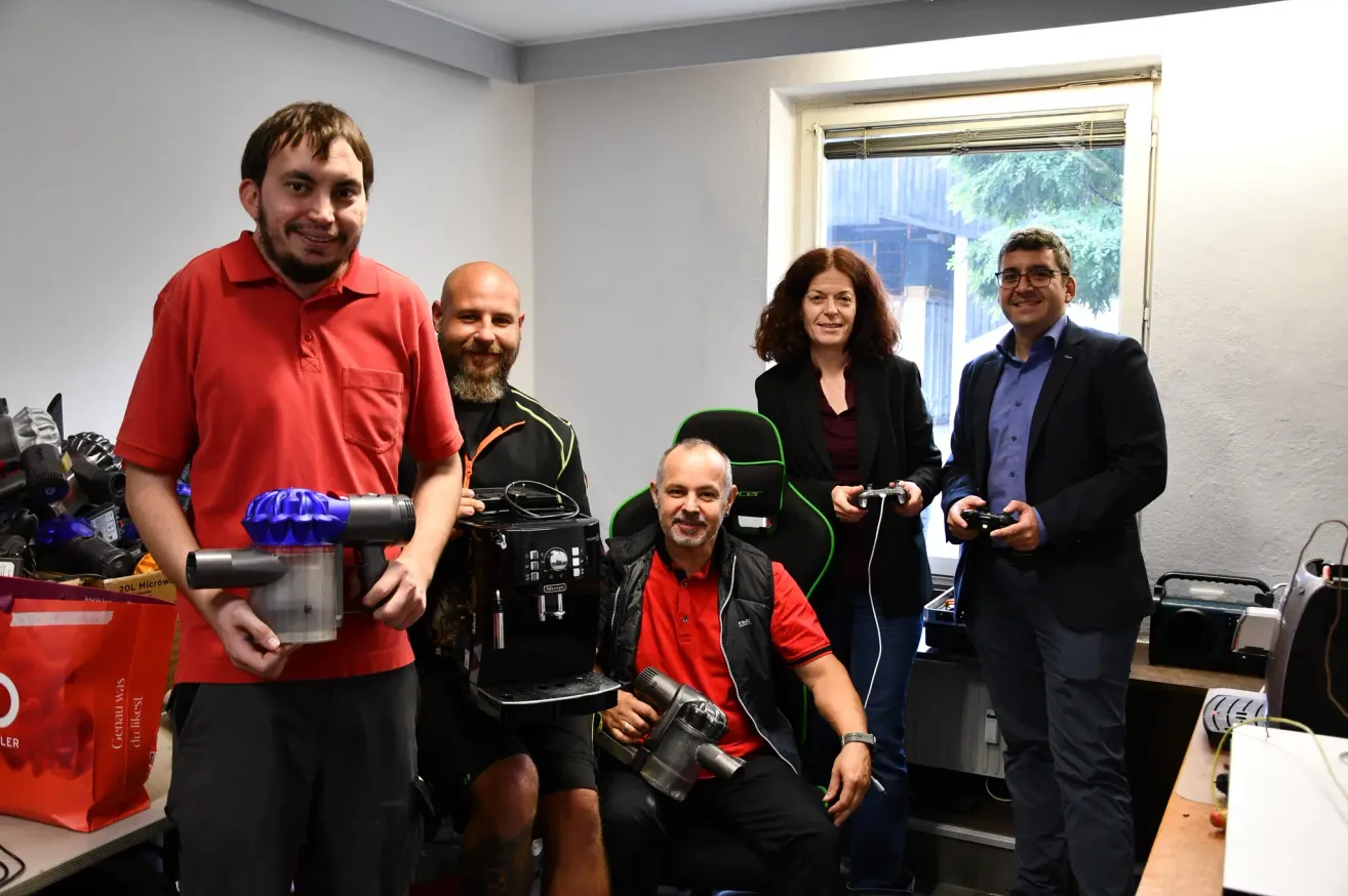 Eine Gruppe von Menschen ist in einem Raum und hält Staubsauger und Controller in der Hand. Ein Mann sitzt, ein anderer steht, und zwei Frauen und ein Mann stehen hinter ihnen.