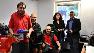 Eine Gruppe von Menschen ist in einem Raum und hält Staubsauger und Controller in der Hand. Ein Mann sitzt, ein anderer steht, und zwei Frauen und ein Mann stehen hinter ihnen.