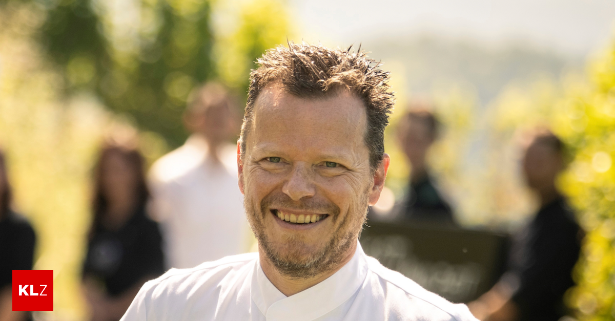 Ein lächelnder Mann in einer weißen Kochuniform steht vor einem unscharfen Hintergrund von Menschen, wahrscheinlich in einem Garten.