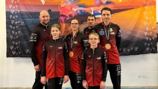 Sechs Personen stehen vor einem Banner mit dem Text RDX und IKW World Cup. Sie tragen alle rote und schwarze Uniformen und haben Medaillen um den Hals.