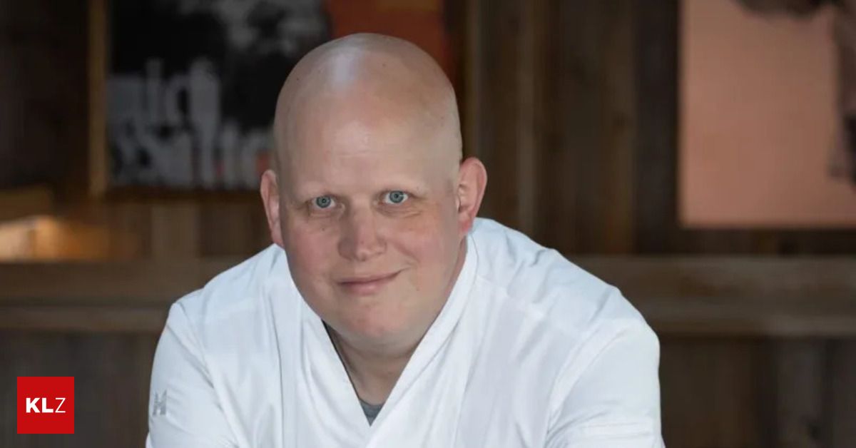 Ein kahlköpfiger Mann in einem weißen Gewand lächelt für ein Foto. Hinter ihm ist eine hölzerne Wand mit einem schwarzen Rahmen.