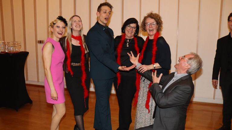 Eine Gruppe von sechs Personen, in festlicher Kleidung gekleidet, posiert für ein Foto, wobei drei Frauen rote Federboas tragen, eine Frau eine Brille trägt und zwei Männer, von denen einer eine Geste macht.