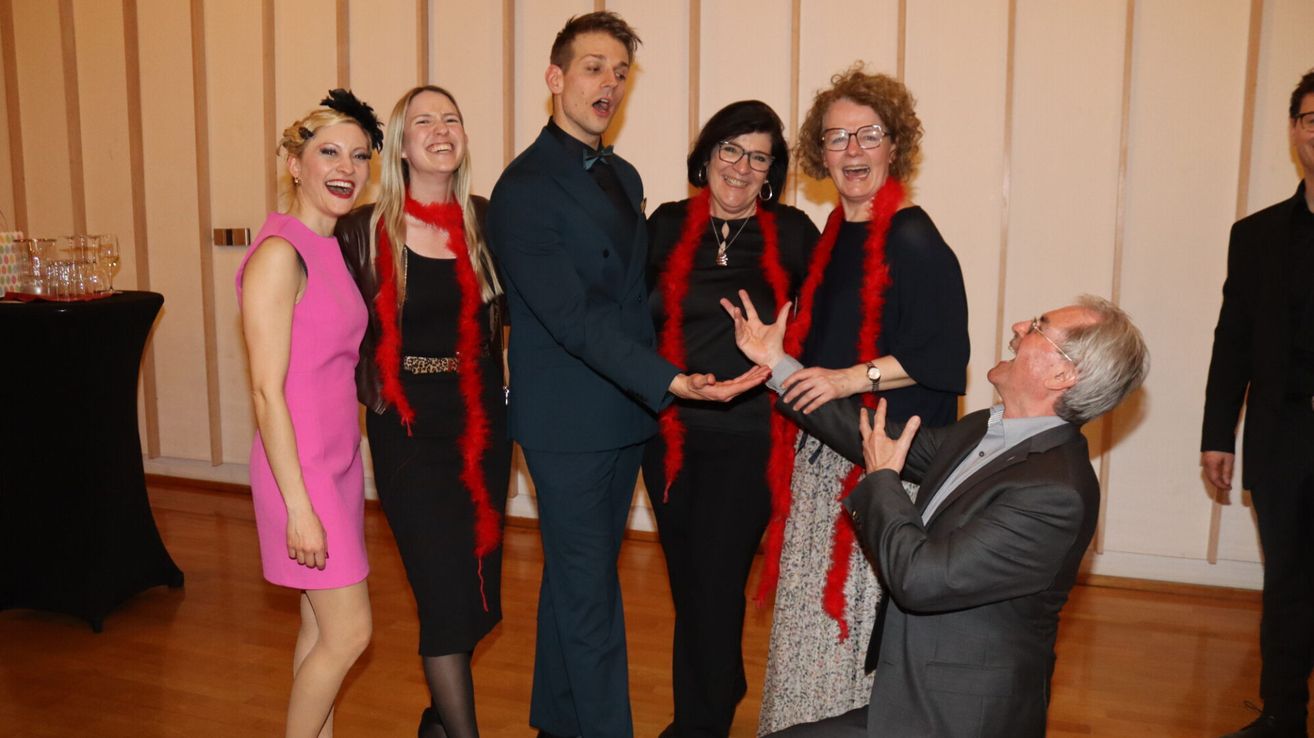 Eine Gruppe von sechs Personen, in festlicher Kleidung gekleidet, posiert für ein Foto, wobei drei Frauen rote Federboas tragen, eine Frau eine Brille trägt und zwei Männer, von denen einer eine Geste macht.