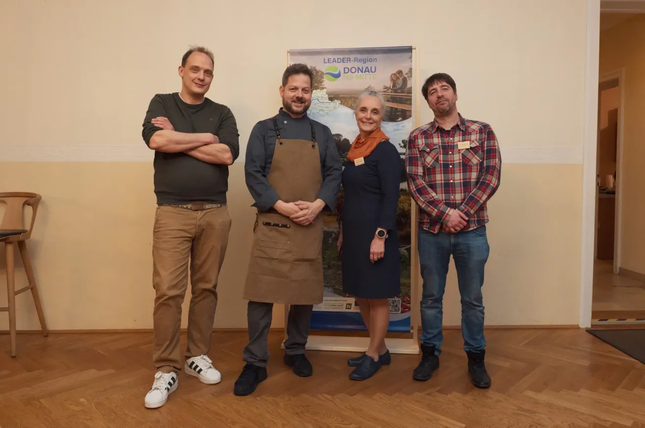 Vier Menschen stehen lächelnd vor einem Banner. Einer trägt eine braune Schürze, ein anderer ein kariertes Hemd und ein Dritter eine Uhr. Die Frau in der Mitte hat eine Schürze um den Hals.