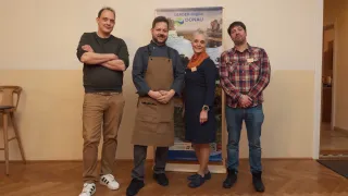 Vier Menschen stehen lächelnd vor einem Banner. Einer trägt eine braune Schürze, ein anderer ein kariertes Hemd und ein Dritter eine Uhr. Die Frau in der Mitte hat eine Schürze um den Hals.