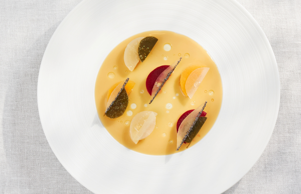 Eine gelbe Suppe in einer weißen Schüssel ist mit Scheiben verschiedener Gemüse und Kräutern garniert und sitzt auf einer hellen Oberfläche.